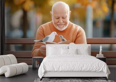 An older man is sitting on a bench reading a book while two birds are perched on his shoulder Wall mural