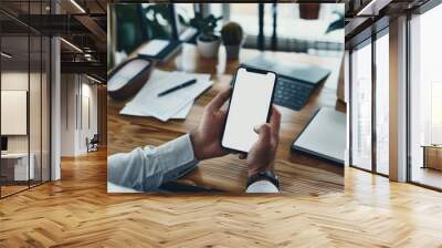 Mockup of a man's hands holding smart phone with blank white screen while sitting at the wooden table in modern office Wall mural