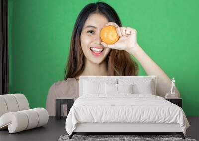 Funny cheerful young Asian woman holding two oranges on her eyes over green isolated background. Wall mural