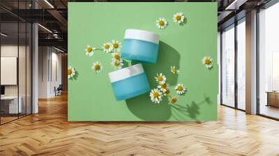 Concept for advertising cosmetics with natural ingredient - chamomilla. Two blue cream jars with flowers displayed on a green background. Chamomilla helps promote cell renewal for healthy glowing skin Wall mural