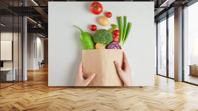 The photo shows a brown supermarket paper bag containing vegetables, held by two hands and dumped on a white table top, was shot from top angle. Space for advertising Wall mural