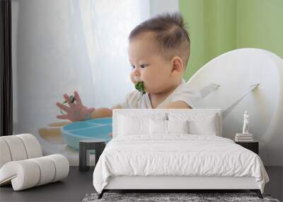 The baby is sitting in a high chair and learning to eat green vegetables Wall mural