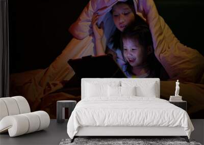 Happy lovely family. two cute Asian kids in pajamas watching cartoon movie from tablet under blanket together in bedroom at night Wall mural