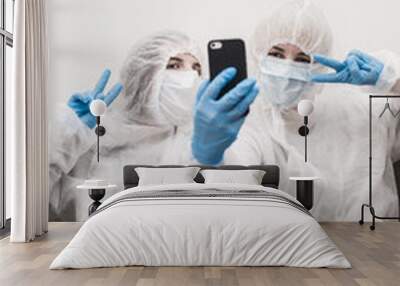 Two people in protective white translucent suit, blue rubber gloves, medical mask are sitting on sofa home and taking selfie on smartphone. Stay home during the coronavirus pandemic. Wall mural