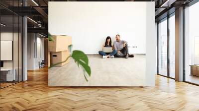 Man sitting with wife using laptop on hardwood floor against wall in new unfurnished home Wall mural