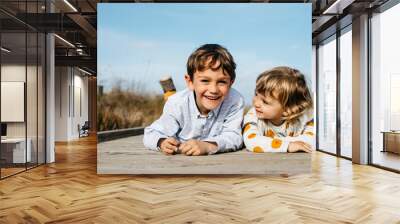 Portrait of boy and his little sister lying side by side on boardwalk having fun Wall mural