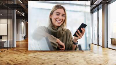 Portrait of happy young woman sitting in waiting area with cell phone Wall mural