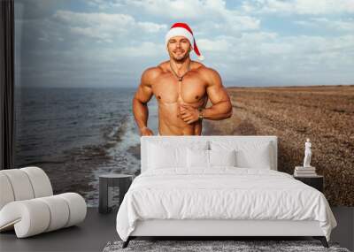 Smiling muscular young man wearing Santa hat running at beach against cloudy sky Wall mural