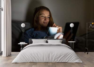 Young woman lying in bed at home with cup of tea at night using tablet Wall mural