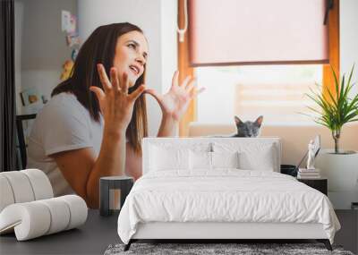 Young woman in white t-shirt sitting with a cat on her lap at the wooden table at home with laptop and notebook, working Wall mural