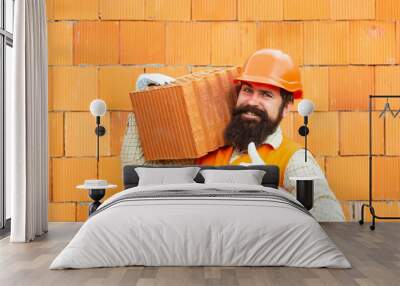 Builder shows thumb up. Labor protection at a construction site. Safety rules for builders. Happy builder in orange helmet holds brick against the background of the wall. Wall mural