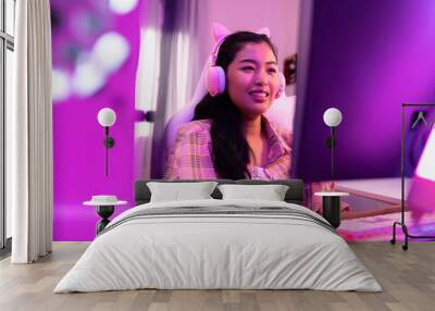 Excited and smiling gamer girl in cute headset with a mic playing an online video game and live streaming. Young Asian woman talking to players and audience on computer in neon led light room at home Wall mural