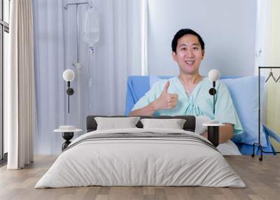 Portrait of a smiling Asian young male patient sitting on hospital bed room showing thumbs up gesture and sign to camera Wall mural