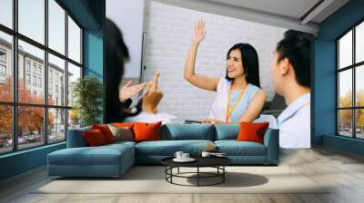 Young smiling Asian business woman raising hand up in the air and asking question in corporate meeting room. Audience is curious in active discussion Wall mural