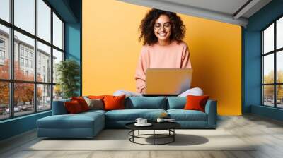 Beautiful young smiling caucasian woman in casual outfit and trendy glasses sitting on the floor isolated on a bright colored background and working on her laptop Wall mural