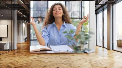 Calm beautiful businesswoman practicing yoga at work, meditating in office with eyes closed. Collegue is on the background. Wall mural