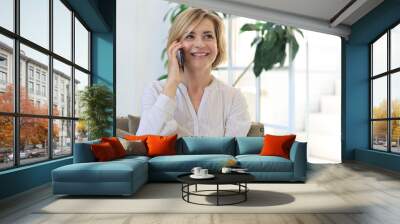 Pretty blond middle aged woman talking on mobile phone sitting on sofa. Wall mural