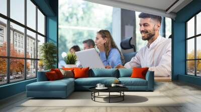 Smiling male call-center operator with headphones sitting at modern office with collegues on the background, consulting online. Wall mural