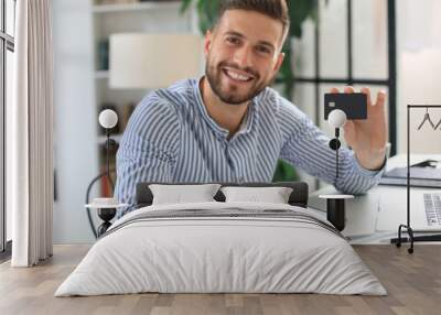 Smiling man sitting in office and pays by credit card with his laptop. Wall mural