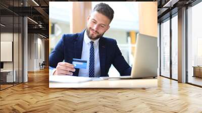 Smiling man sitting in office and pays by credit card with his laptop. Wall mural