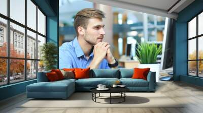 Young man sitting and looking at computer monitor while working in office. Wall mural