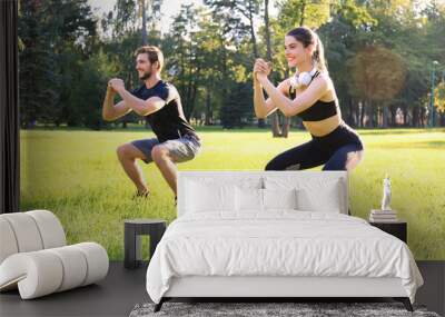 Young sporty man and woman doing workout and squatting together in green park during summer sunny day. Wall mural