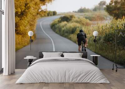 Back view of strong male cyclist with athletic body shape riding bike at the paved road among trees and green bushes in black protective helmet and sportswear. Concept of training  outdoors Wall mural