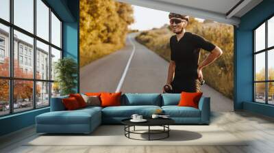 Cheerful bearded man in activewear, black helmet and sport glasses sitting on bike and looking on camera with beautiful nature around. Concept of active and healthy lifestyle. Wall mural