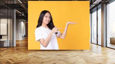 Beautiful Asian woman wearing casual clothes smile and pointing finger to an empty palm isolated on yellow background studio portrait. Wall mural