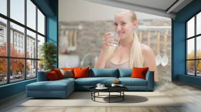 A beautiful European woman smiles happily, holding a glass of water, drinking natural mineral water from a clear glass, inside a residential. A healthy woman shows her face, complete physical health. Wall mural