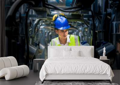 A focused engineer wearing a safety vest and helmet inspects machinery in an industrial setting. The professional diligently checks equipment to ensure proper functionality and safety standards. Wall mural