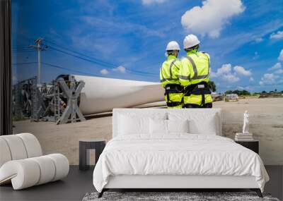 Engineers in high visibility gear assess a large wind turbine blade at a construction site under a bright blue sky, representing the implementation of renewable energy technology. Wall mural