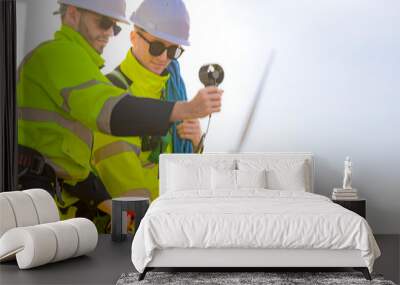 Two engineers wearing high visibility safety jackets and helmets measure wind speed with an anemometer and analyze data on a tablet at a renewable energy site. The cloudy sky adds depth to the scene. Wall mural