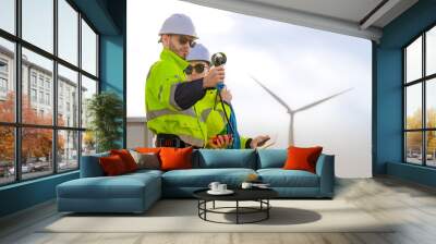 Two engineers wearing high visibility safety jackets and helmets measure wind speed with an anemometer and analyze data on a tablet at a renewable energy site. The cloudy sky adds depth to the scene. Wall mural