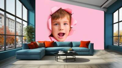 Happy child boy shows face in paper hole over pink background with blank space. Effect of torn paper.  Wall mural