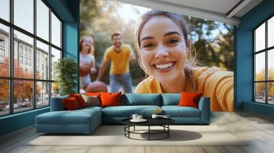 Young woman taking a selfie with friends playing basketball in a sunny park, all smiling and having fun. Wall mural