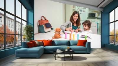 A woman and a child are sitting at a table with a book and a bag Wall mural