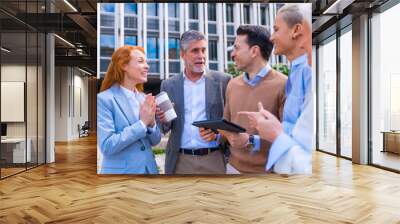 Cheerful group of coworkers laughing and looking at a tablet outdoors in a corporate office area, businesswoman and businessman and executives in commercial area Wall mural