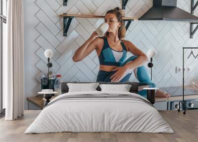 Athletic woman having breakfast in the kitchen, girl sitting on table and drinking Wall mural