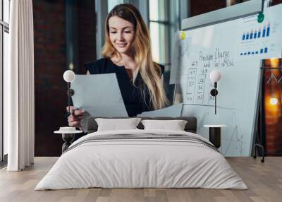 Female office worker working on her presentation standing near white board reading the report printed on paper Wall mural