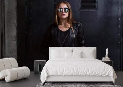 Half-length portrait of sexy young woman wearing sunglasses, black swimsuit and leather jacket posing over dark background Wall mural