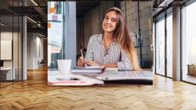 portrait of smiling female student writing looking at camera sitting at table Wall mural