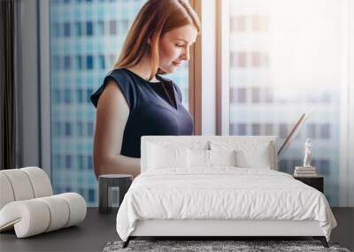 Portrait of young woman working holding laptop standing against panoramic window with city view Wall mural