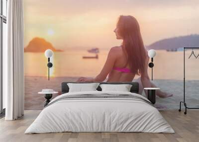 Rear view of slim female model sitting on seashore wearing bikini looking away during sunrise with sun path reflected  water Wall mural