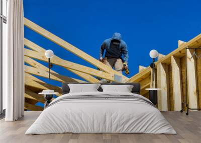 Builder on the unfinished roof with nailing wooden beams in frame house under construction with aerial vie Wall mural