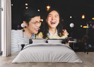 Asian mother and daughter laughing and smiling on a selfie or photo album, using smartphone together at restaurant or cafe, with copy space. Family love, holiday activity, or modern lifestyle concept Wall mural