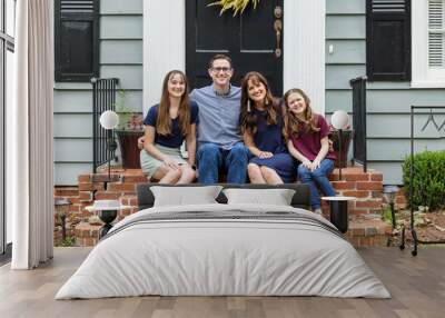 A family with a mother, father, and two daughters sitting outside on the brick porch of a small blue cottage house Wall mural