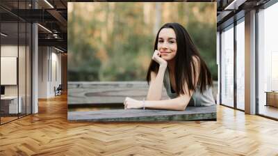 A tall and thin brunette teenager standing and leaning on a park bridge with her head resting on her hands with a smile and joyful expression Wall mural