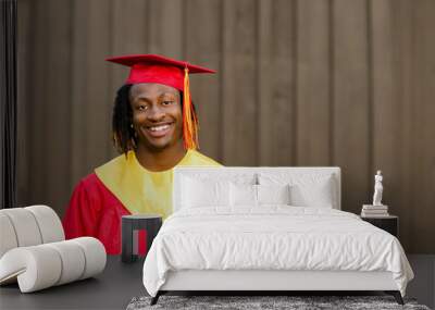 Smiling happy African-American Teen Teenager male Man outside against a brown wall in his red and gold graduation gown Wall mural