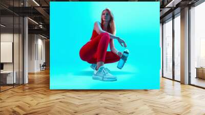 Young beautiful blonde sports girl in red sportswear squatting on a bright neon blue background holding a bottle with a drink in her hand. Wall mural